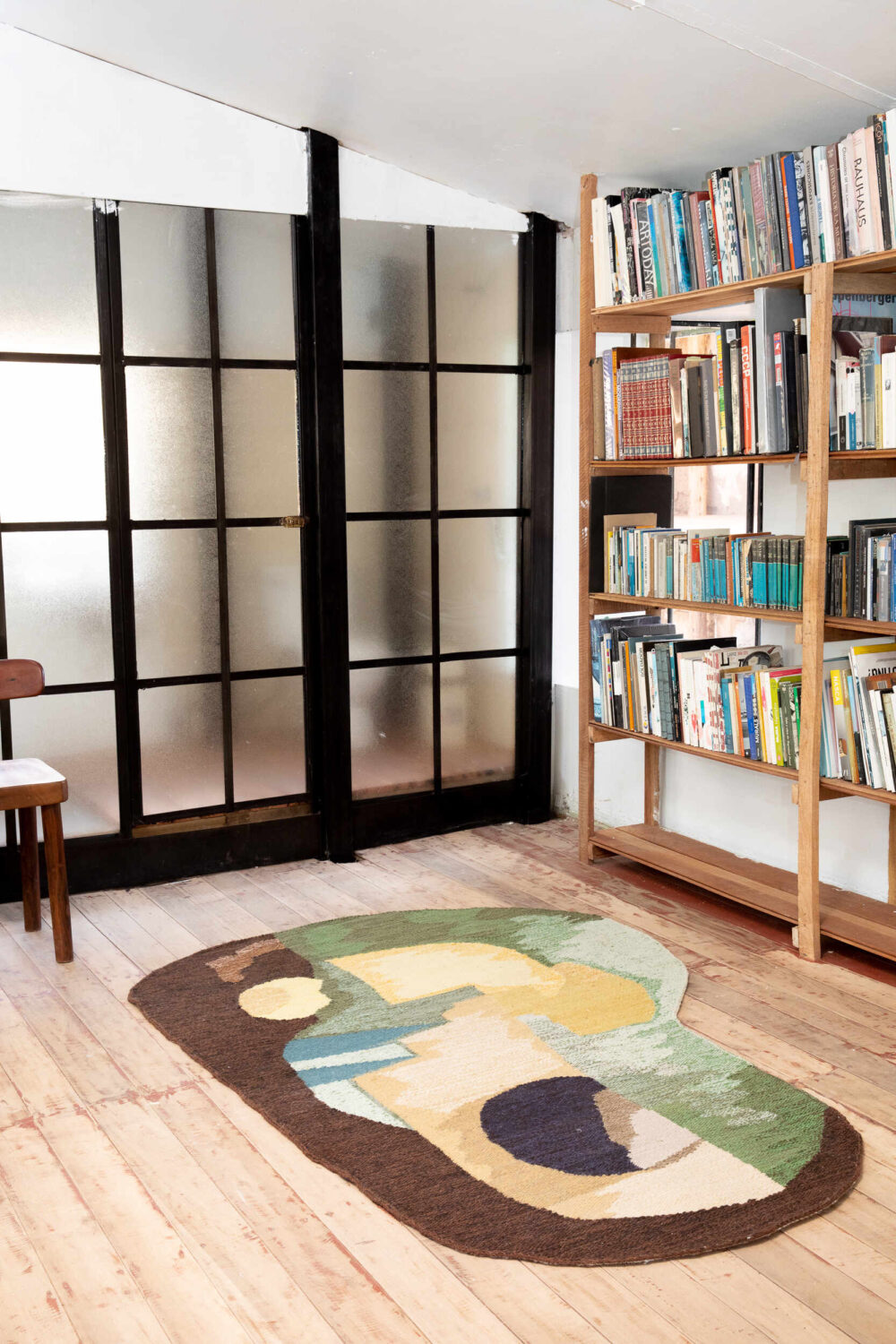 Avocado 01 rug in a wood-floored minimalist space, with thin wood bookshelves full of books and a glass-paneled space divider